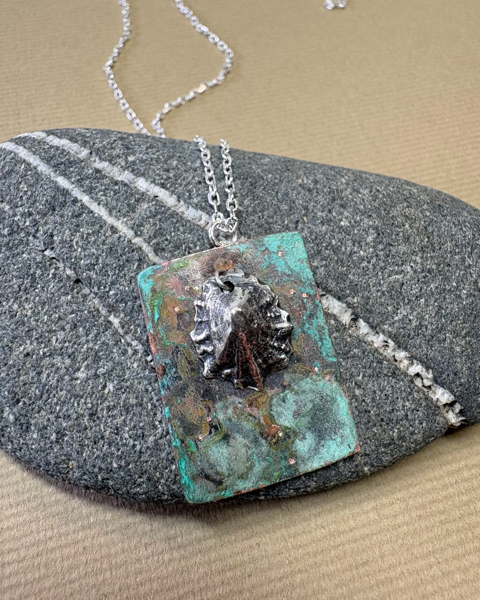 Silver limpet shell in its rock pool with a square pendant featuring an oxidised pattern on copper on a pebble surface