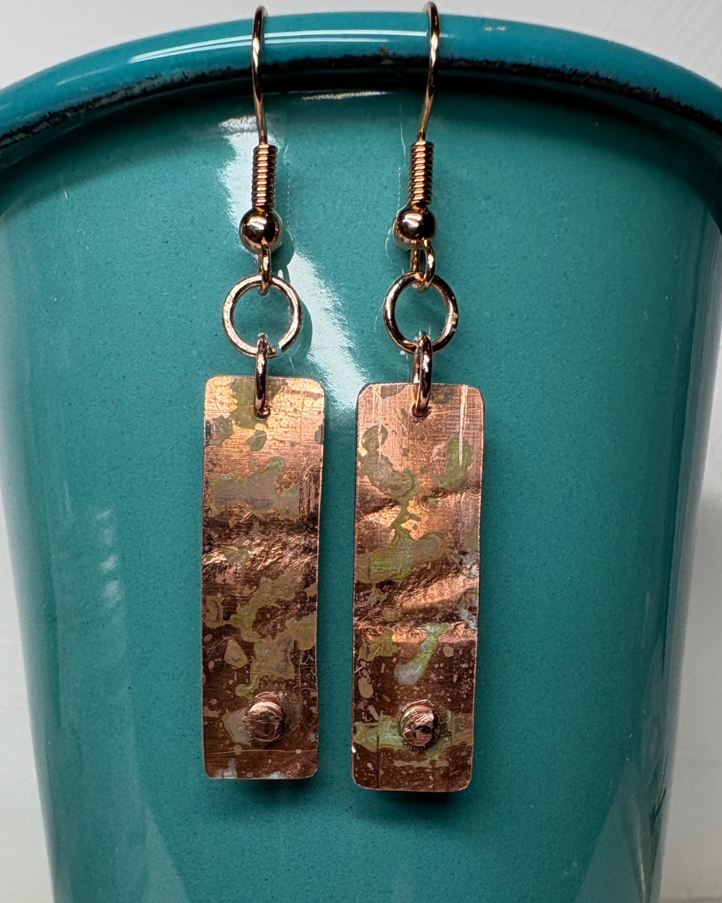 Pair of textured rectangular copper earrings with patina and small raised circle near base on a teal background
