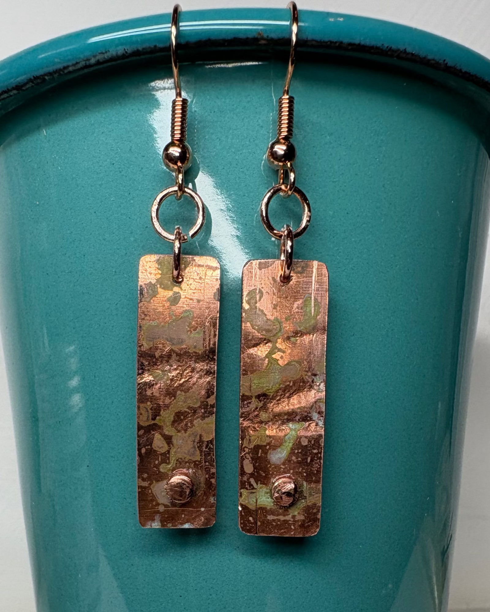 Pair of textured rectangular copper earrings with patina and small raised circle near base on a teal background