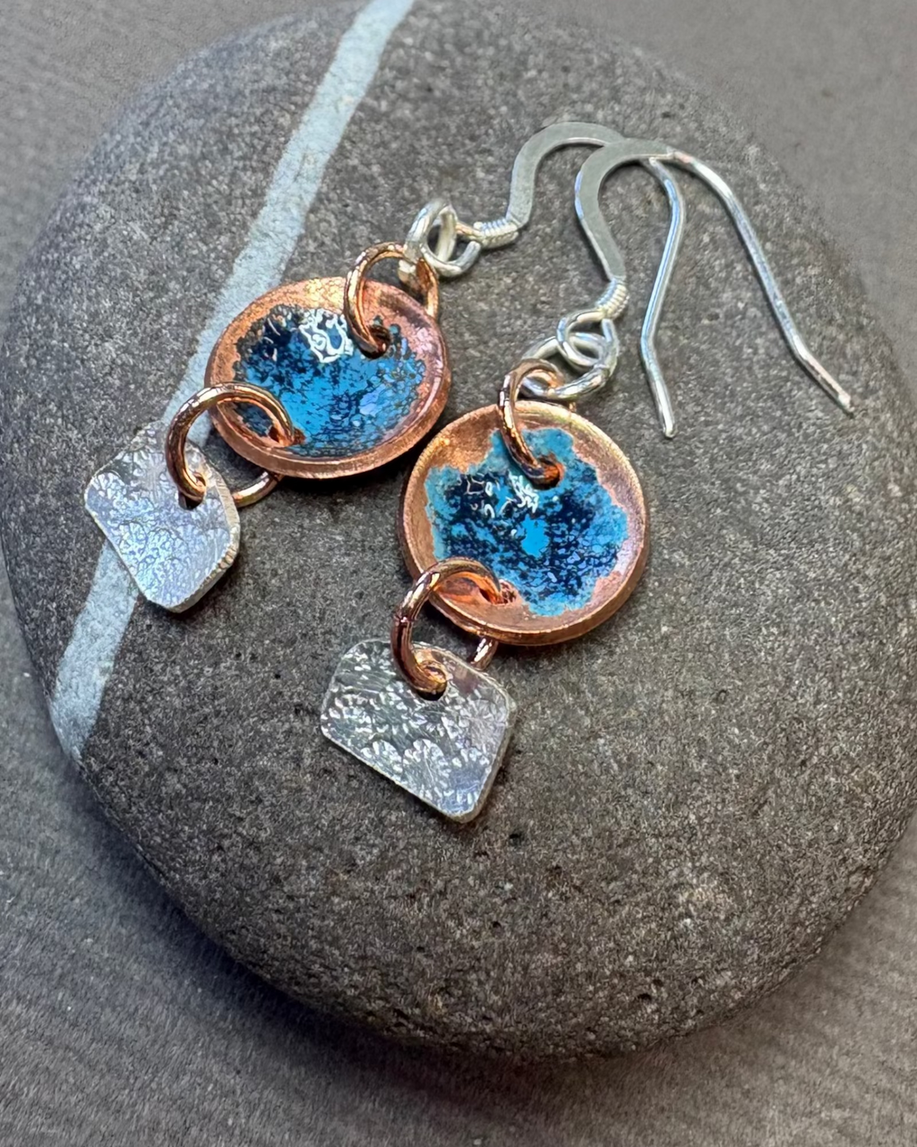 Silver and copper earrings with enamelling on the copper, pictured on a pebble