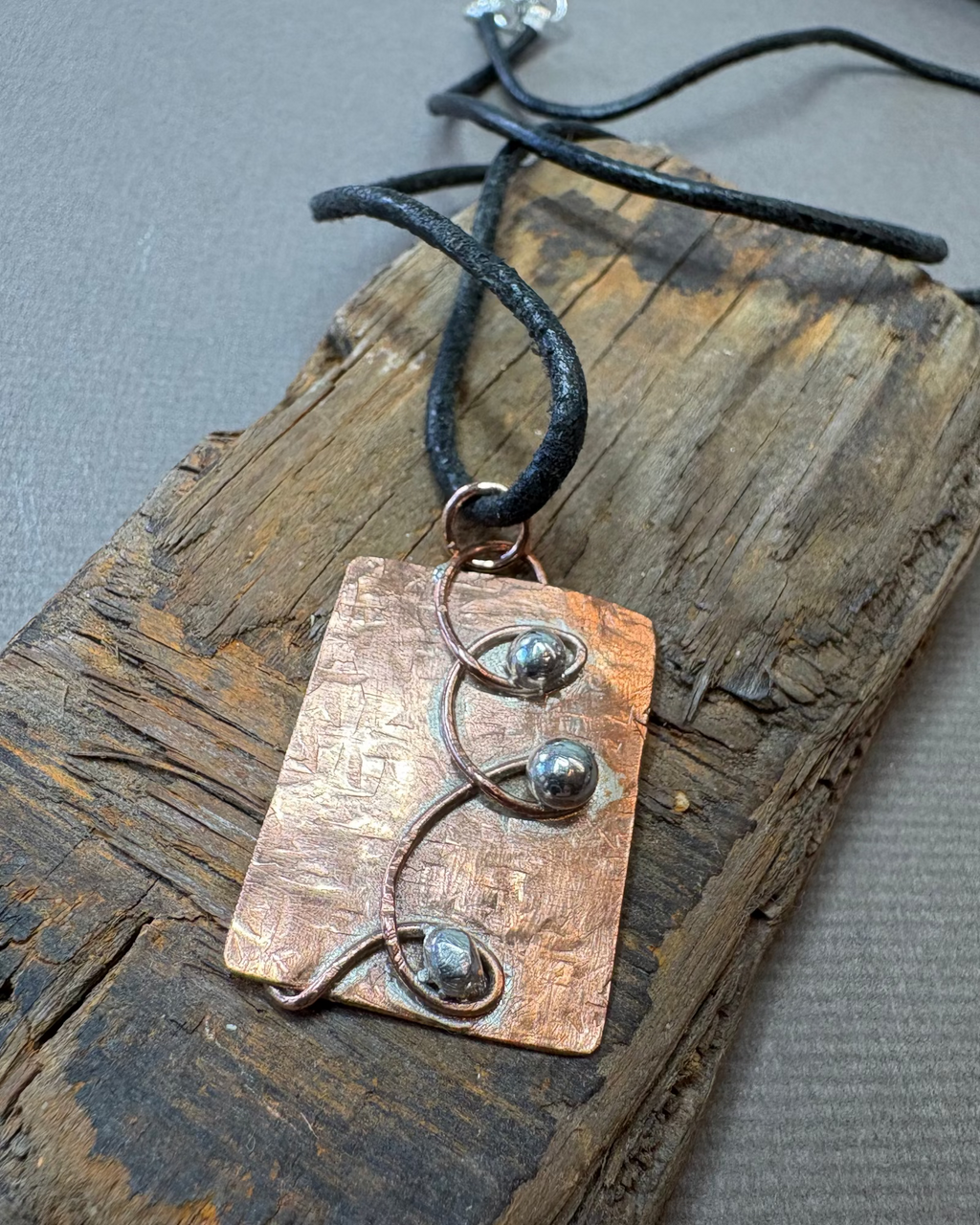 Copper and silver pendant necklace with black leather cord on a driftwood surface