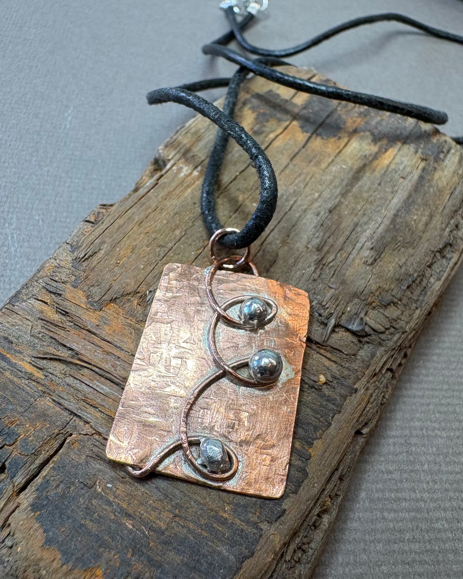 Copper and silver pendant necklace with black leather cord on a driftwood surface