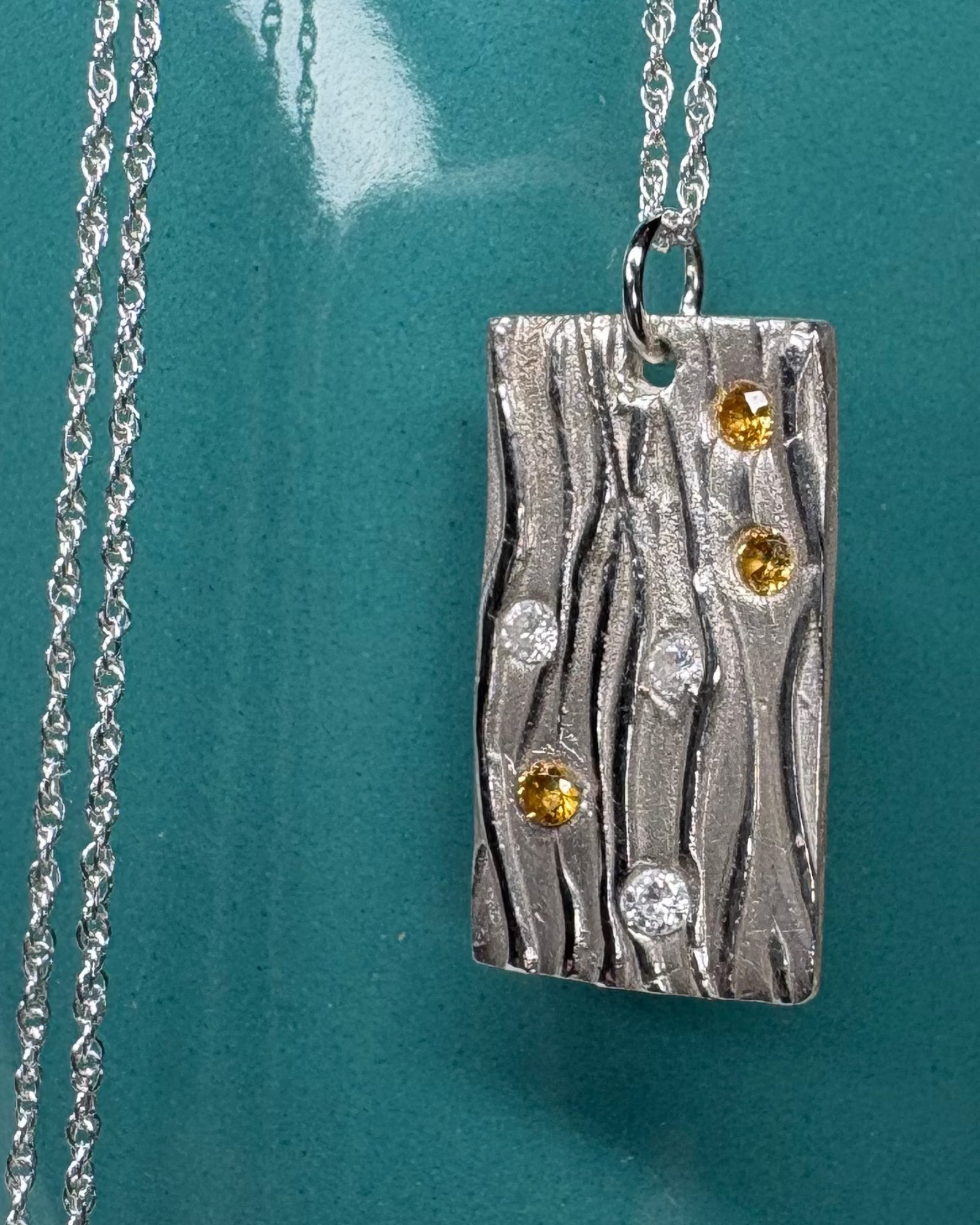 Silver rectangular necklace with a wavy design incorporating gold and white zirconia with a silver chain on a teal background
