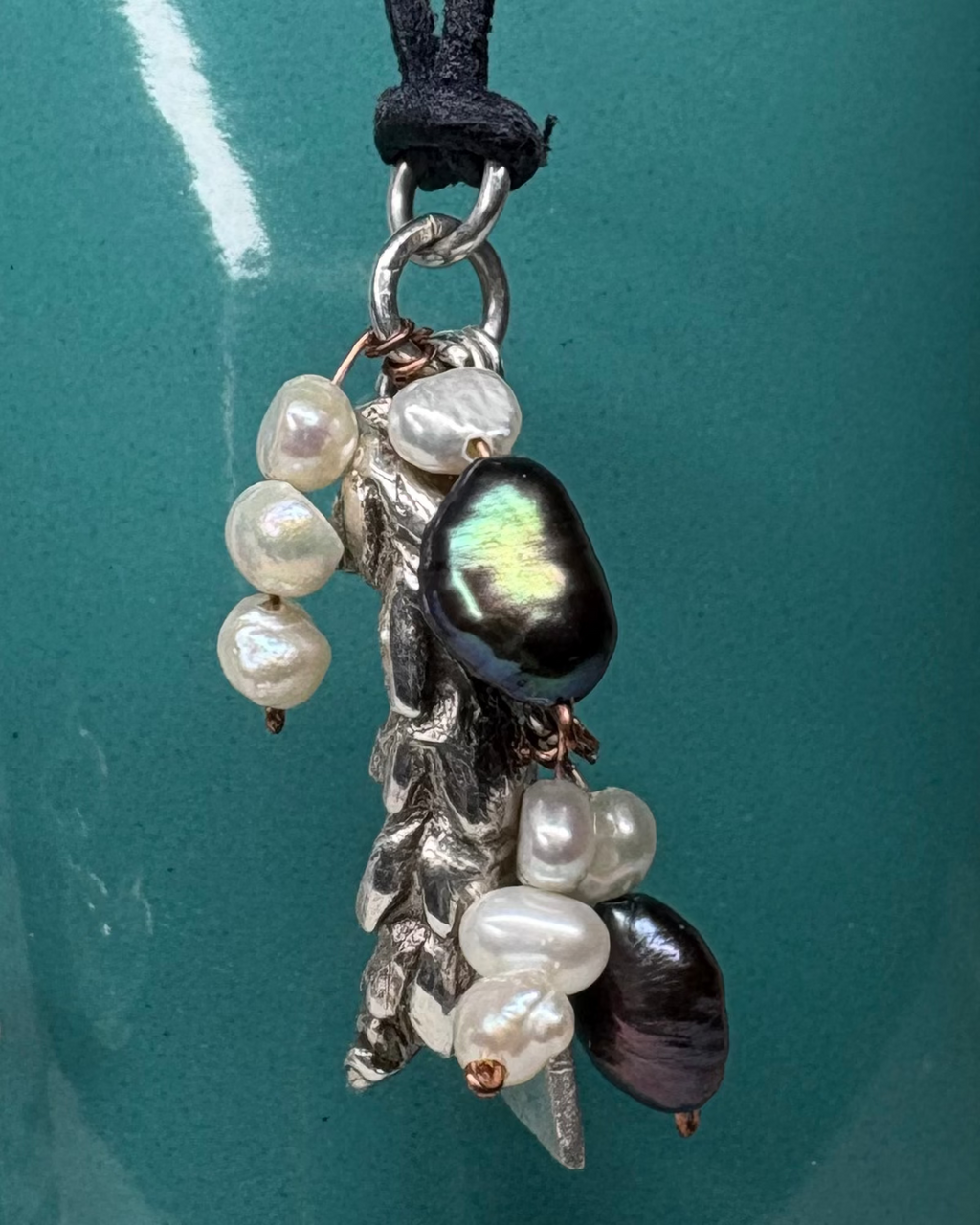 Necklace with fine silver lavender flower charm and freeform peacock and cream pearls with a black leather cord draped over a teal container 