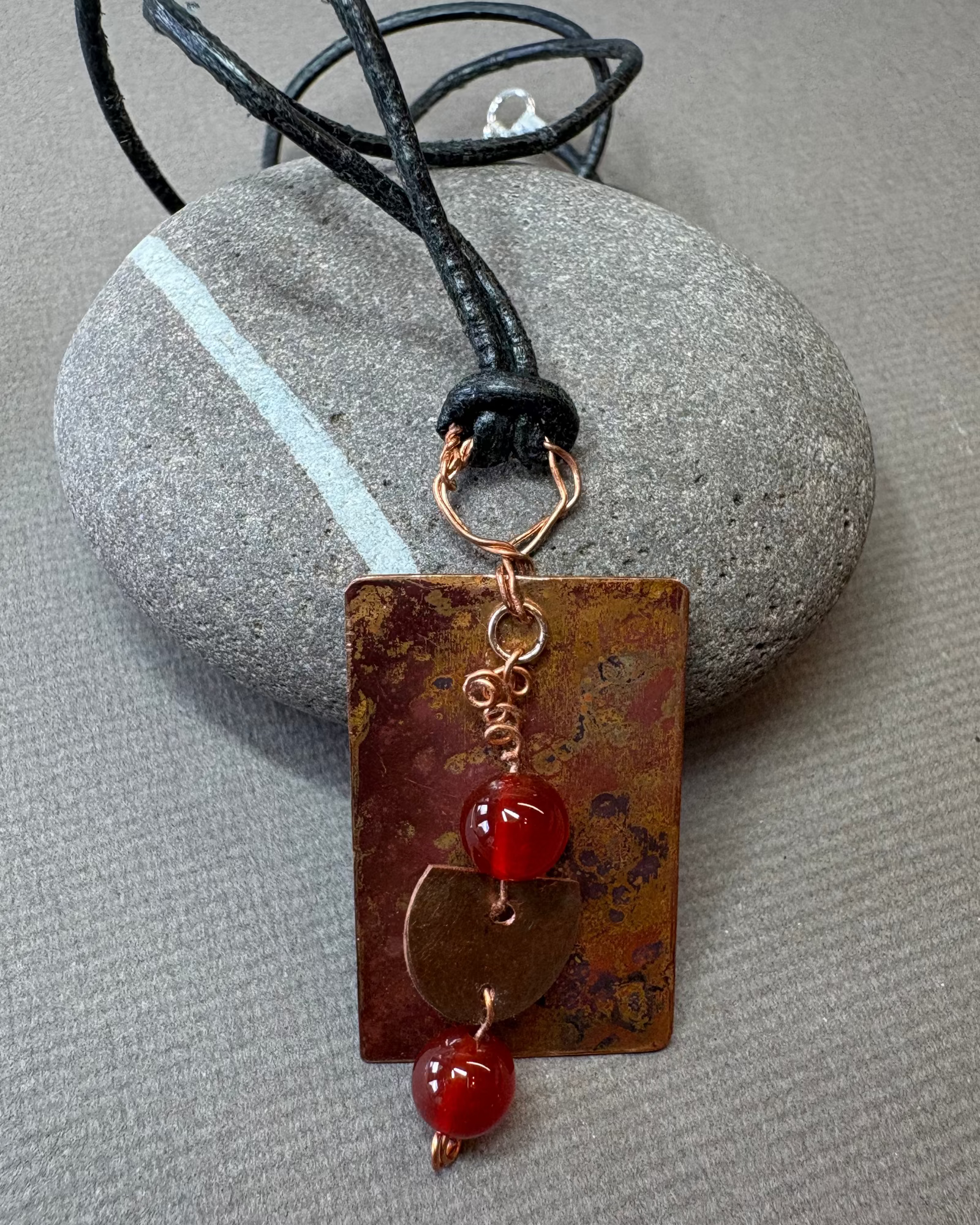 Leather necklace with a rectangular copper oxidised pendant and carnelian beads on a pebble
