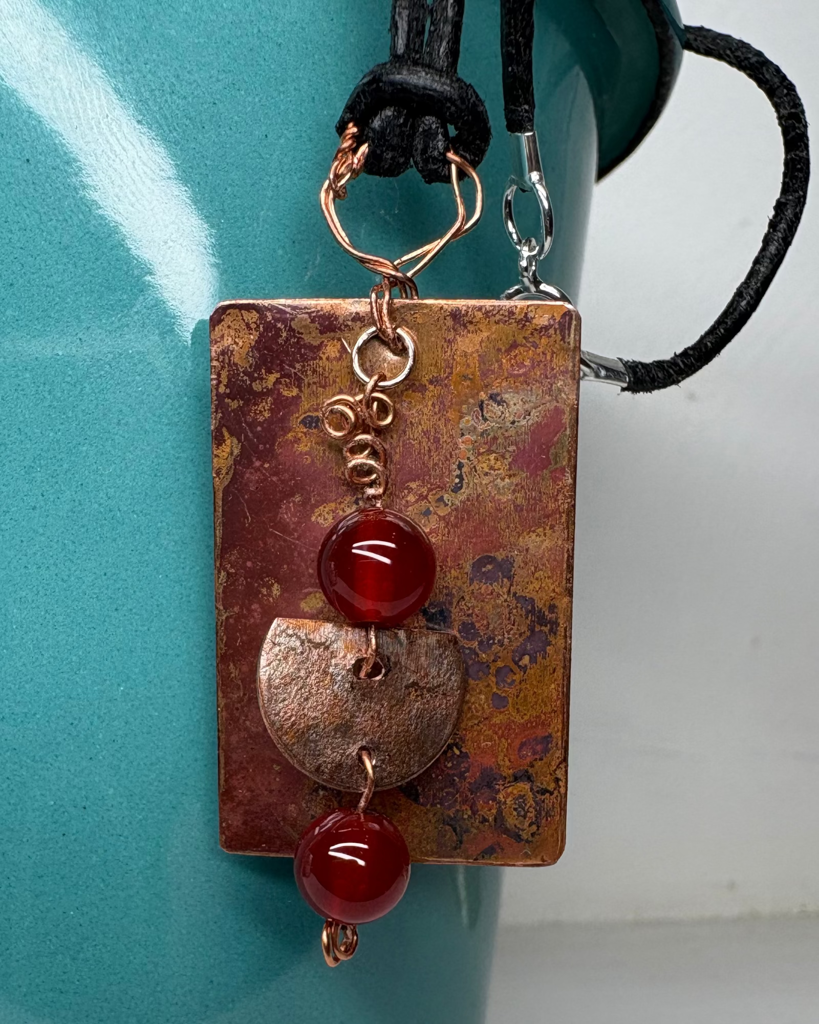 Necklace with a rectangular oxidised copper pendant featuring carnelian beads  and half moon copper charm against a teal mug background