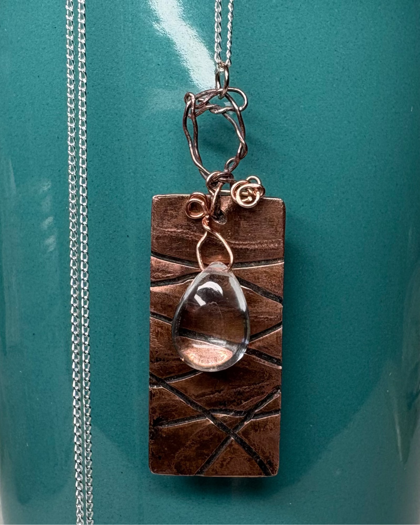 Necklace with rectangular textured copper  with a teardrop quartz stone wired over rectangle hanging on a silver chain pictured on a teal container 