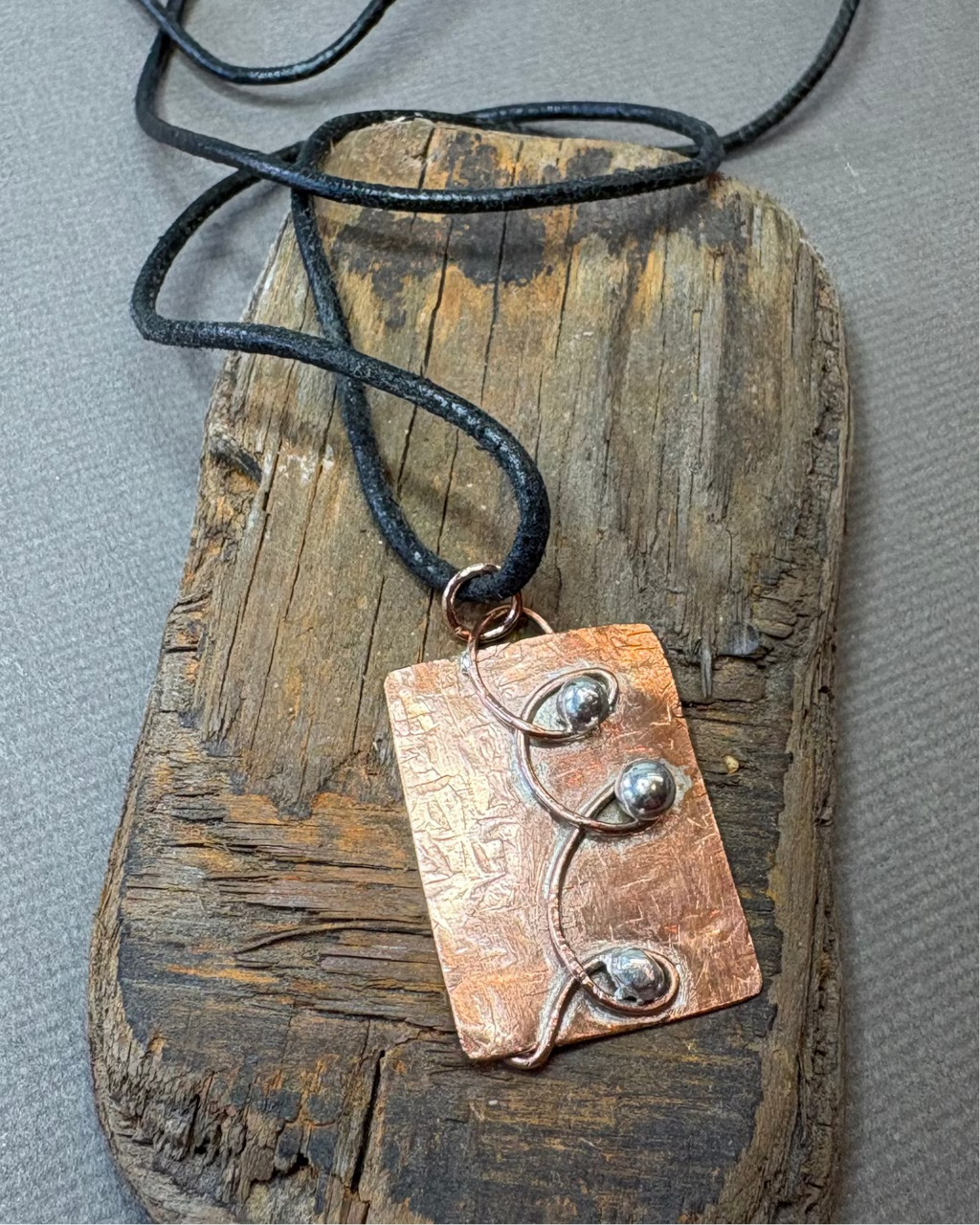 Copper and silver pendant with a black leather cord, placed on a piece of driftwood