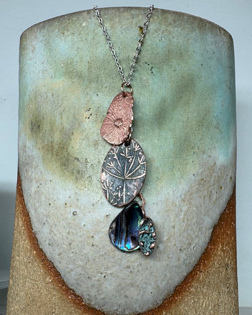 Necklace with textured copper flower head and paua shell charms on a silver chain on a ceramic surface