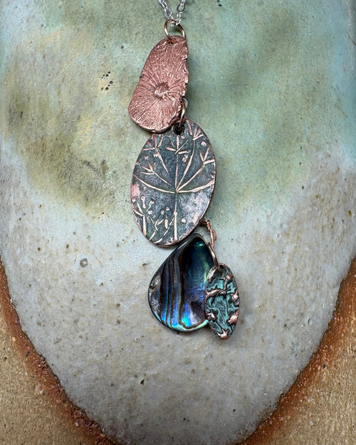 Necklace with textured flower head and paua shell charms on a silver chain on a ceramic surface