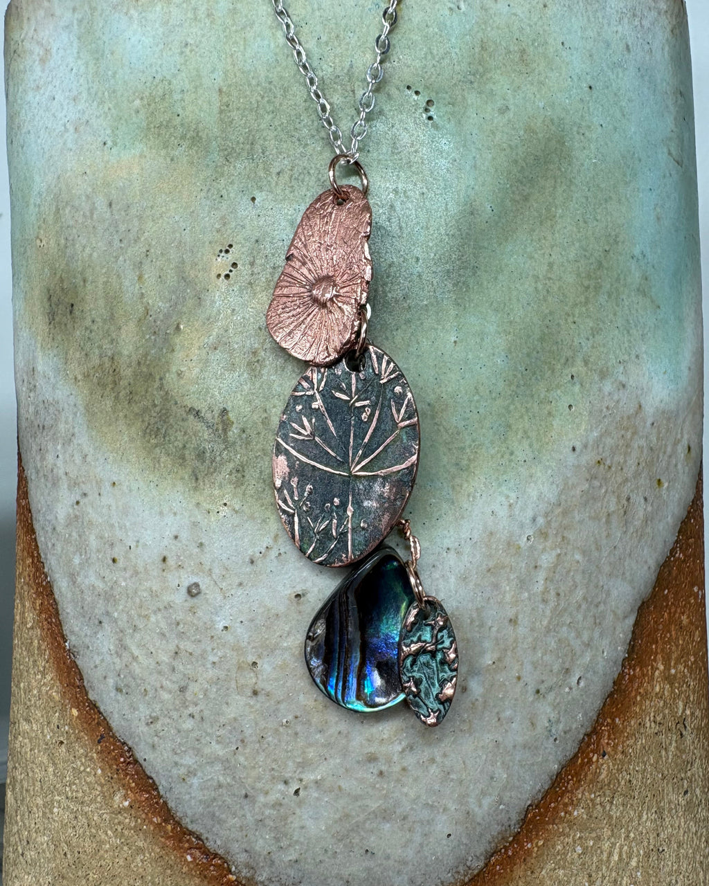 Necklace with textured flower head and paua shell charms on a silver chain on a ceramic surface