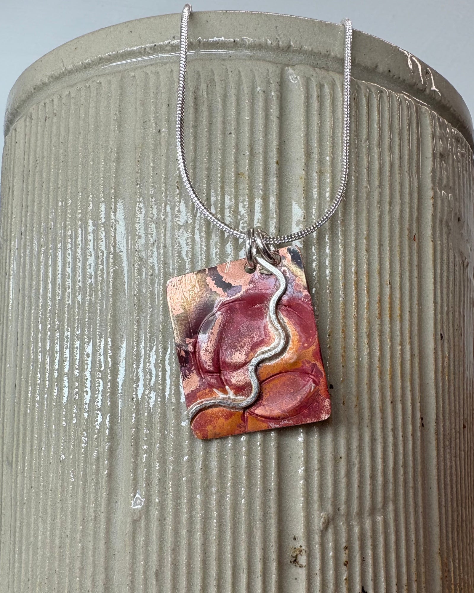A handmade fire oxidised copper pendant necklace with pressed ripples and silver wavy design. This hangs from silver chain on ceramic vase.