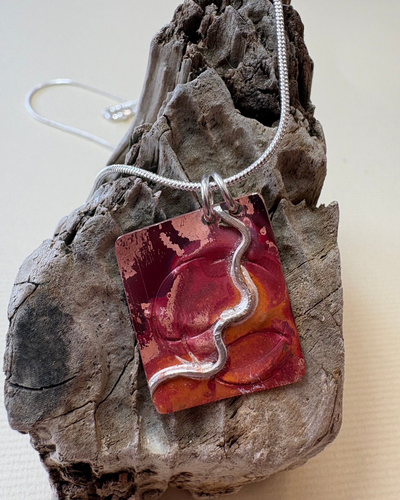 A handmade fire oxidised copper pendant necklace with pressed ripples and silver wavy design. This hangs from silver chain on a piece of driftwood.