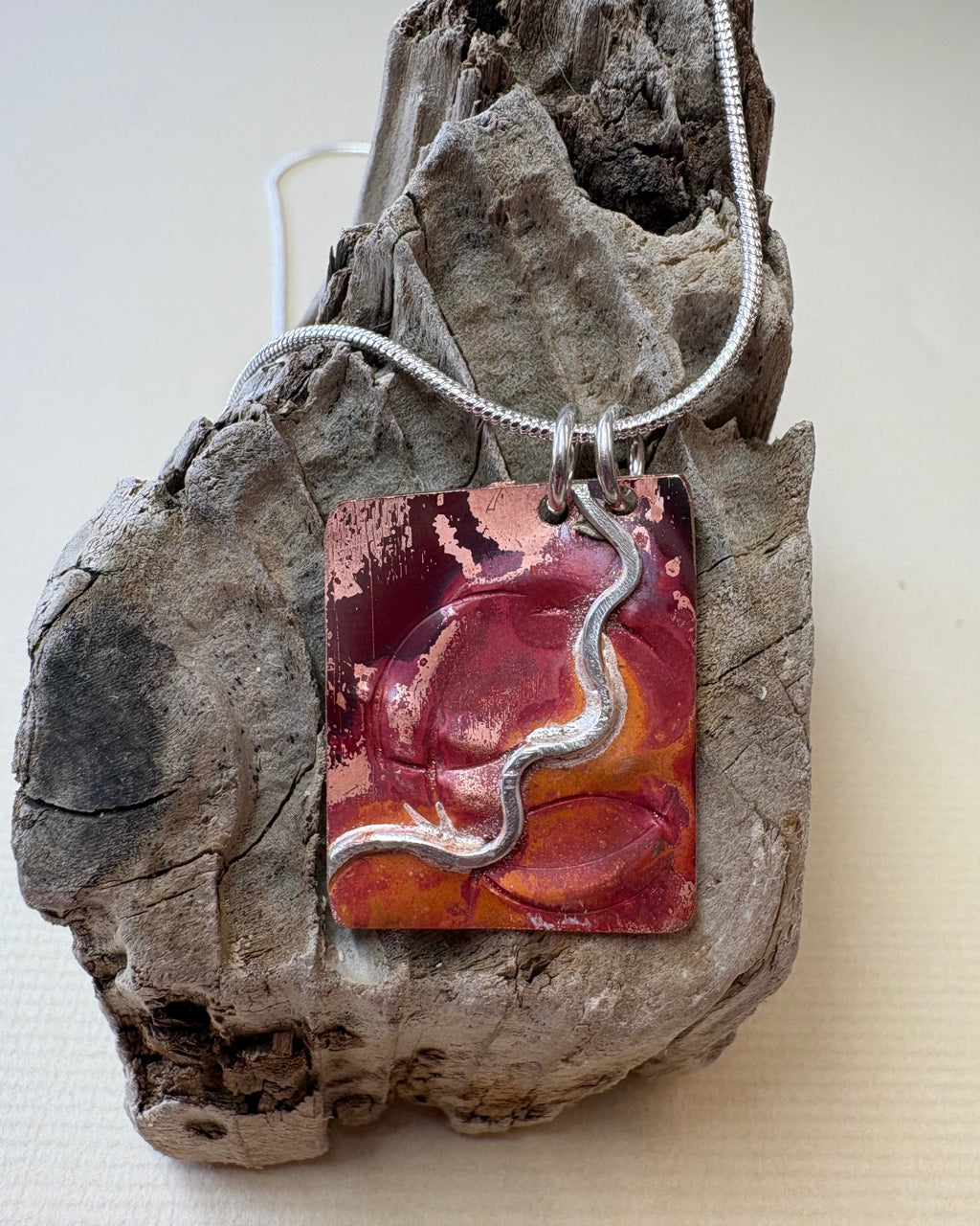 A handmade fire oxidised copper pendant necklace with pressed ripples and silver wavy design. This hangs from silver chain on a piece of driftwood.