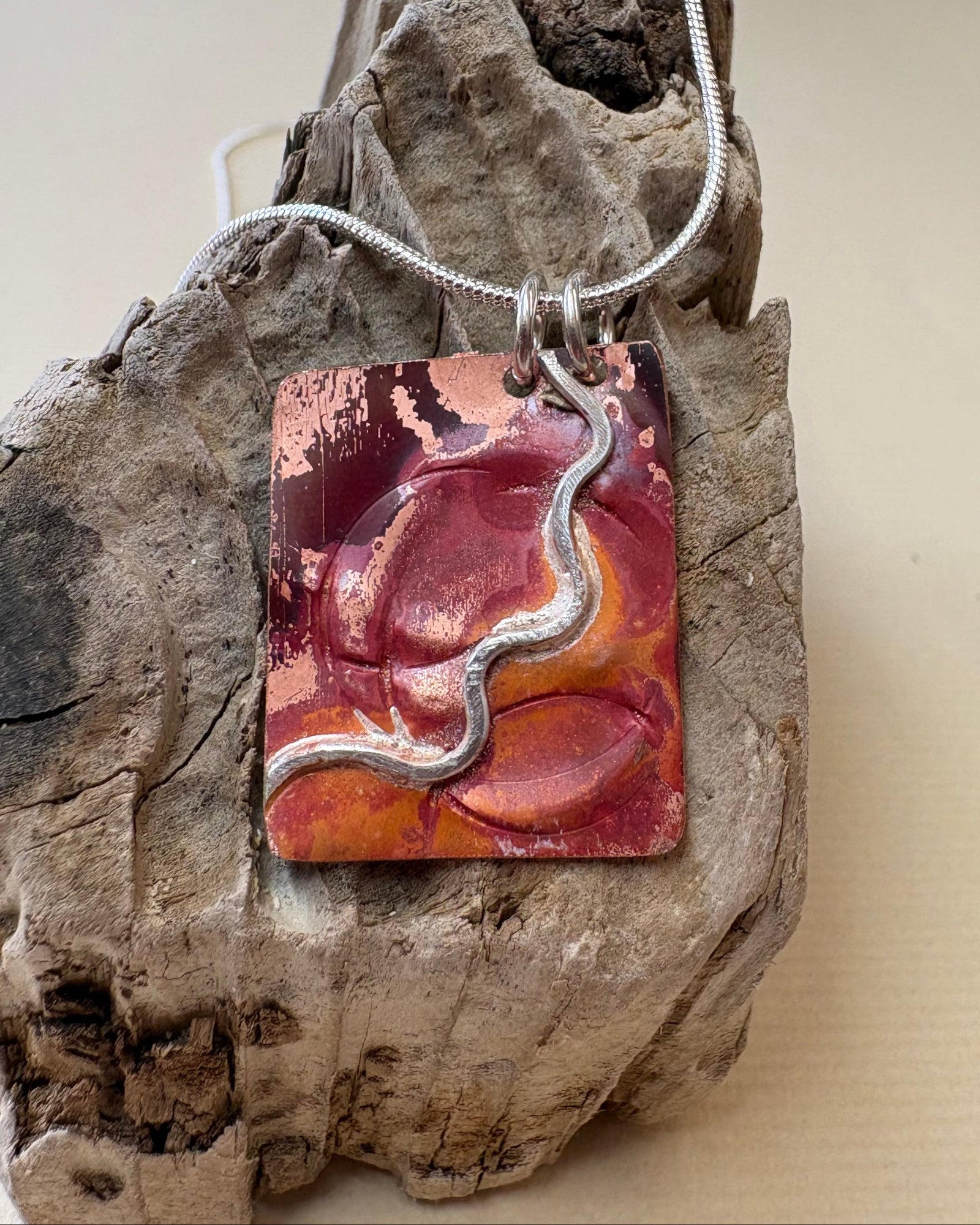 A handmade fire oxidised copper pendant necklace with pressed ripples and silver wavy design. This hangs from silver chain on a piece of driftwood.