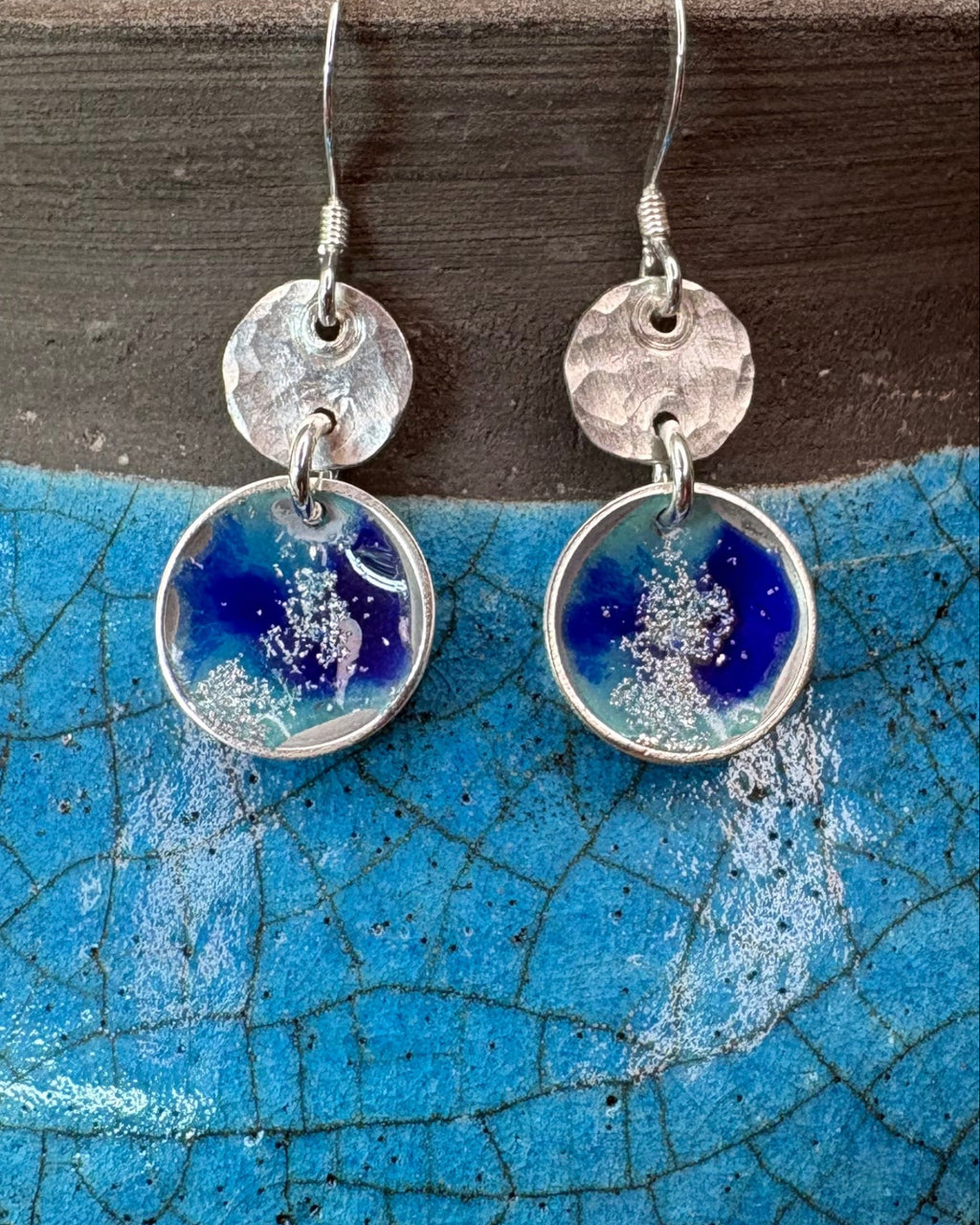 round silver concave enamel disc earrings with blue, aqua and silver to look like pools, with second hammered silver circle disk above, hung on silver ear wires. Pictured on a blue raku vase