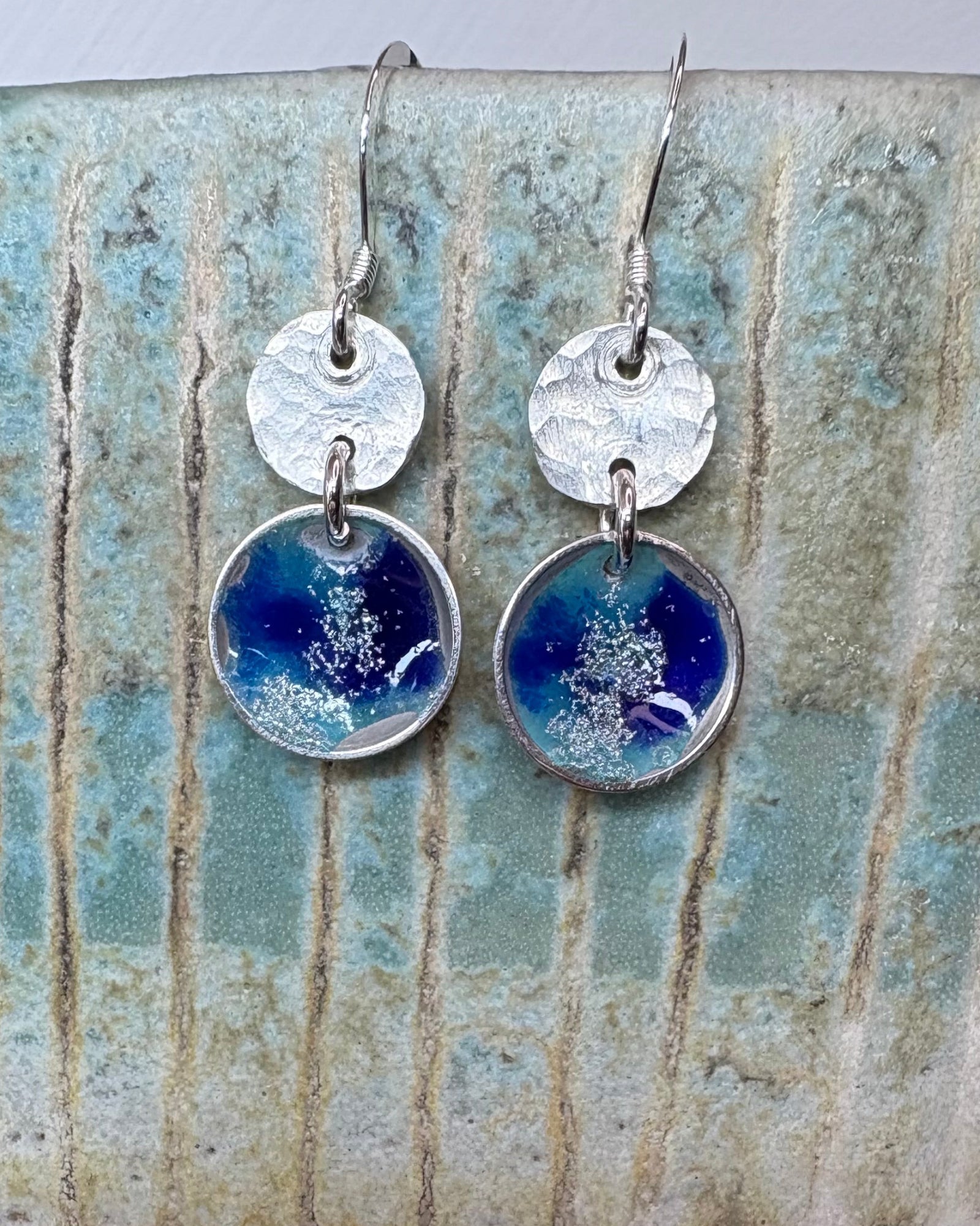 round silver concave enamel disc earrings with blue, aqua and silver to look like pools, with second hammered silver circle disk above, hung on silver ear wires pictured on a ceramic vase