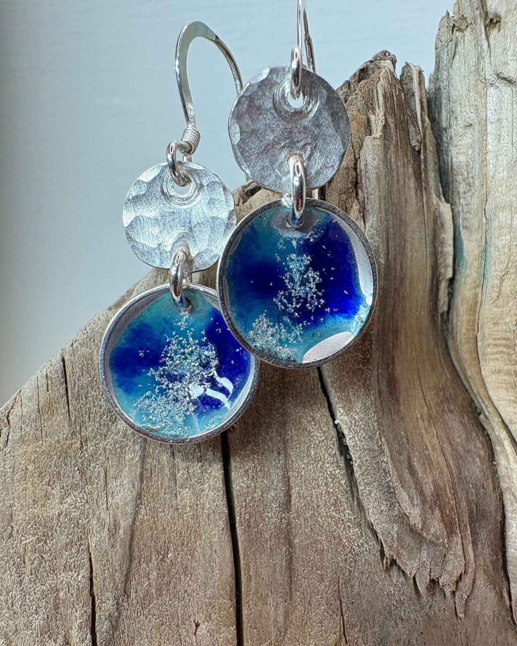 round silver concave enamel disc earrings with blue, aqua and silver to look like pools, with second hammered silver circle disk above, hung on silver ear wires
hanging on a piece of driftwood