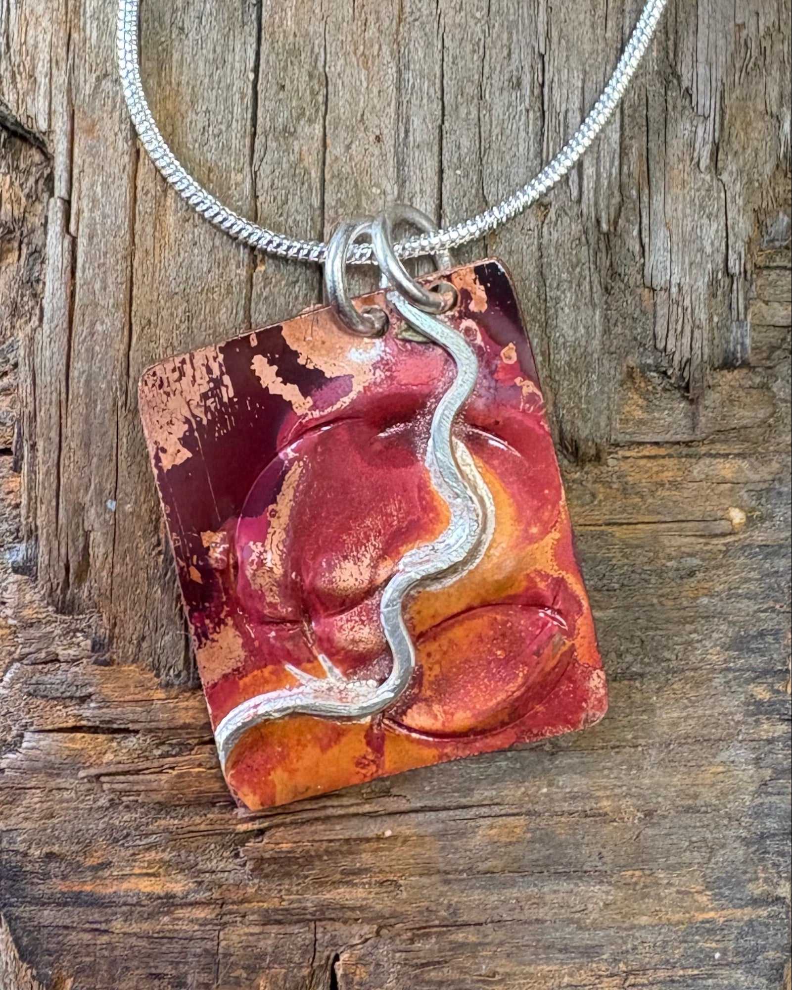 A handmade asymmetric pendant necklace with fire coloured copper with a wave of silver, photographed on a piece of driftwood