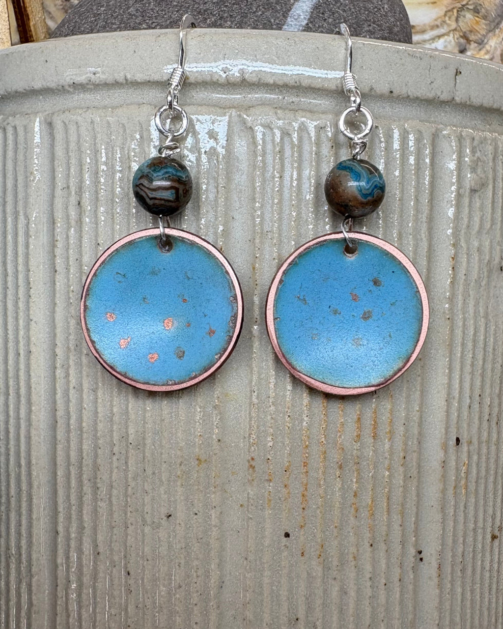 A pair of handmade dangle earrings with round light blue enamel pendants, copper-rimmed edges, and agate beads on silver fishhook wires, displayed on a vintage stoneware pot