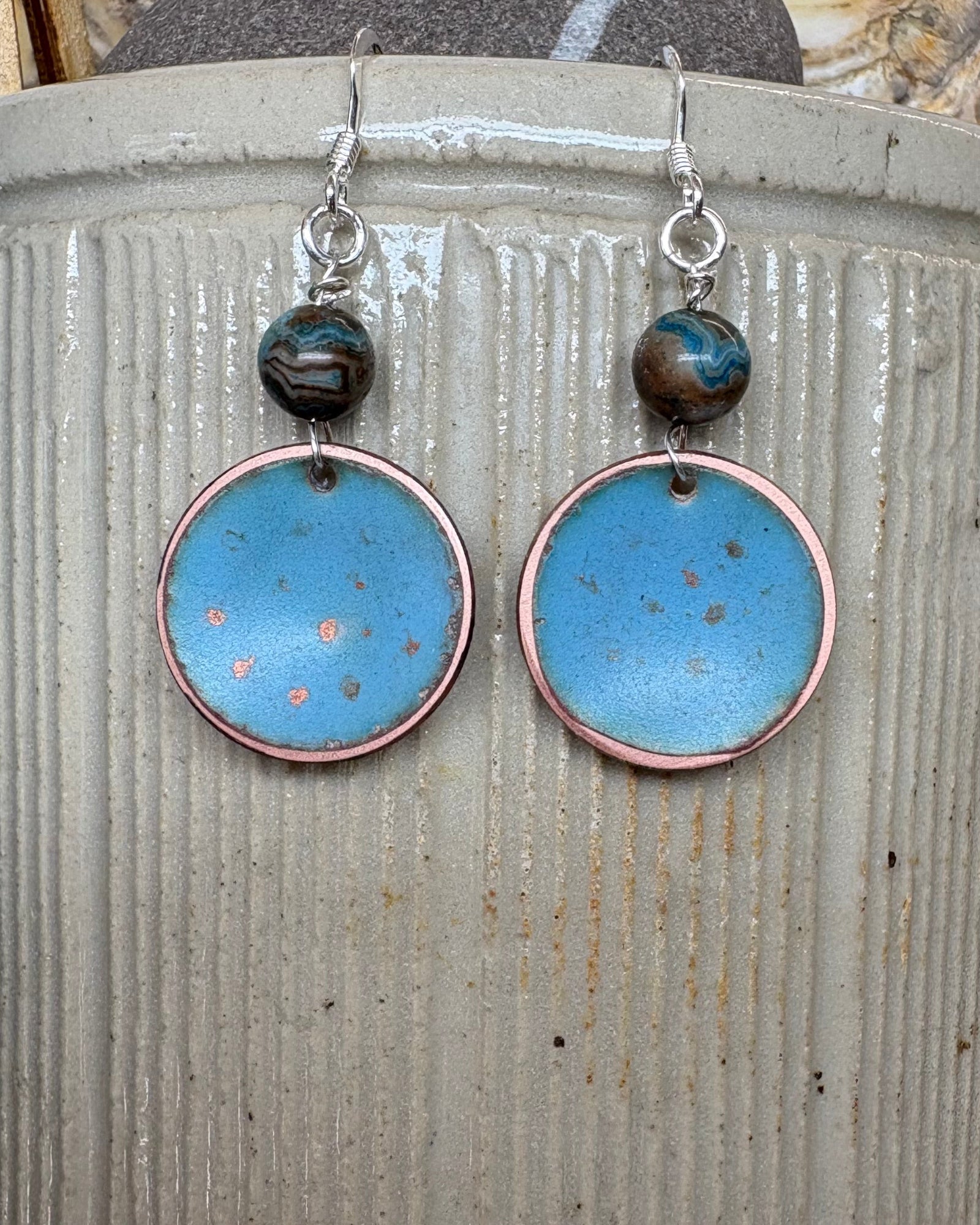 A pair of handmade dangle earrings with round light blue enamel pendants, copper-rimmed edges, and agate beads on silver fishhook wires, displayed on a vintage stoneware pot