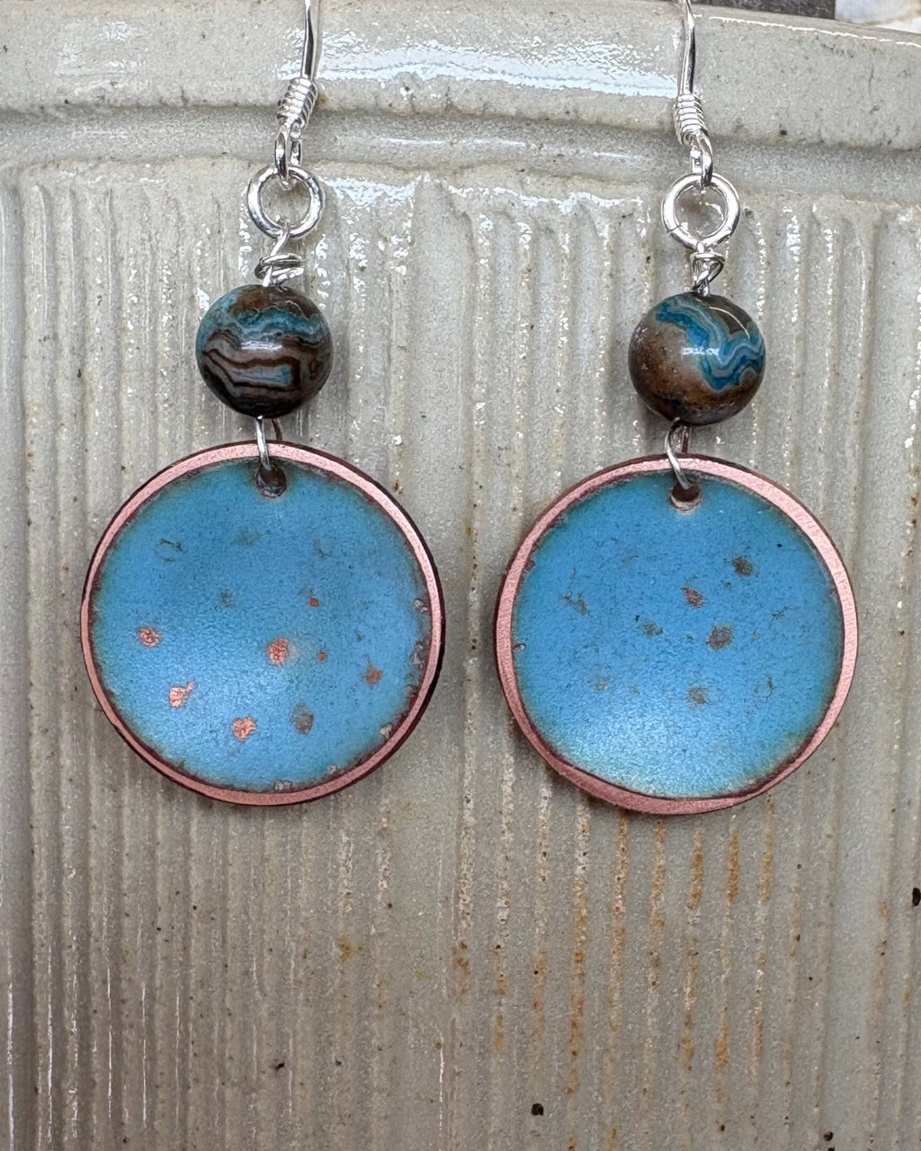 A pair of handmade dangle earrings with round light blue enamel pendants, copper-rimmed edges, and agate beads on silver fishhook wires, displayed on a vintage stoneware pot