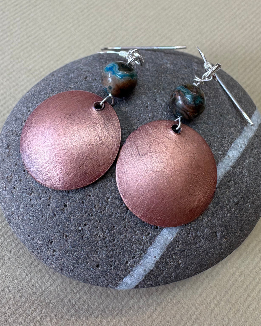 A the back of a pair of handmade dangle earrings brushed copper finish, and agate beads on silver fishhook wires, displayed on a pebble