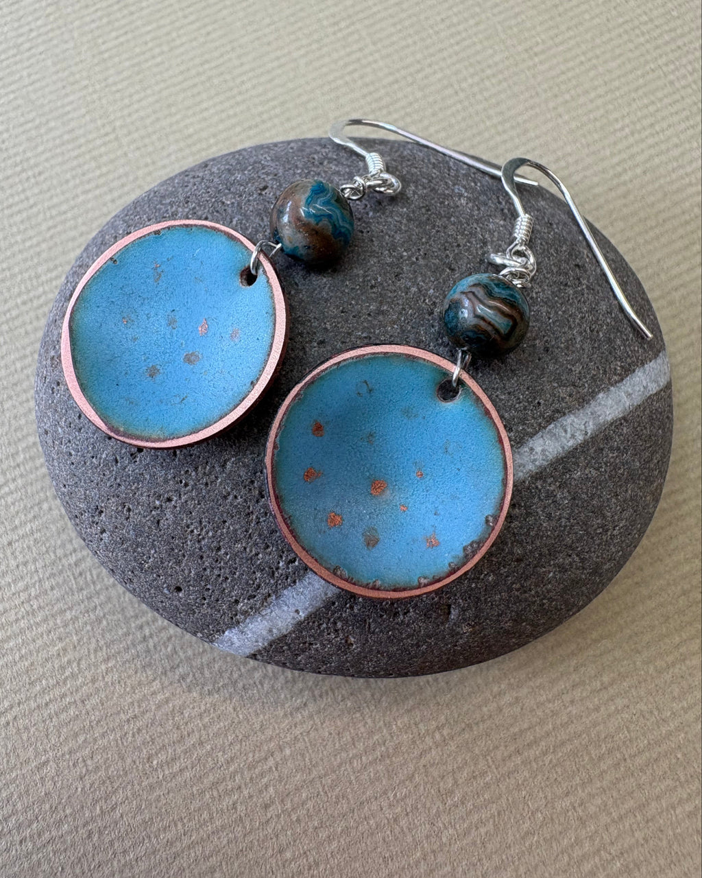A pair of handmade dangle earrings with round light blue enamel pendants, copper-rimmed edges, and agate beads on silver fishhook wires, displayed on a pebble