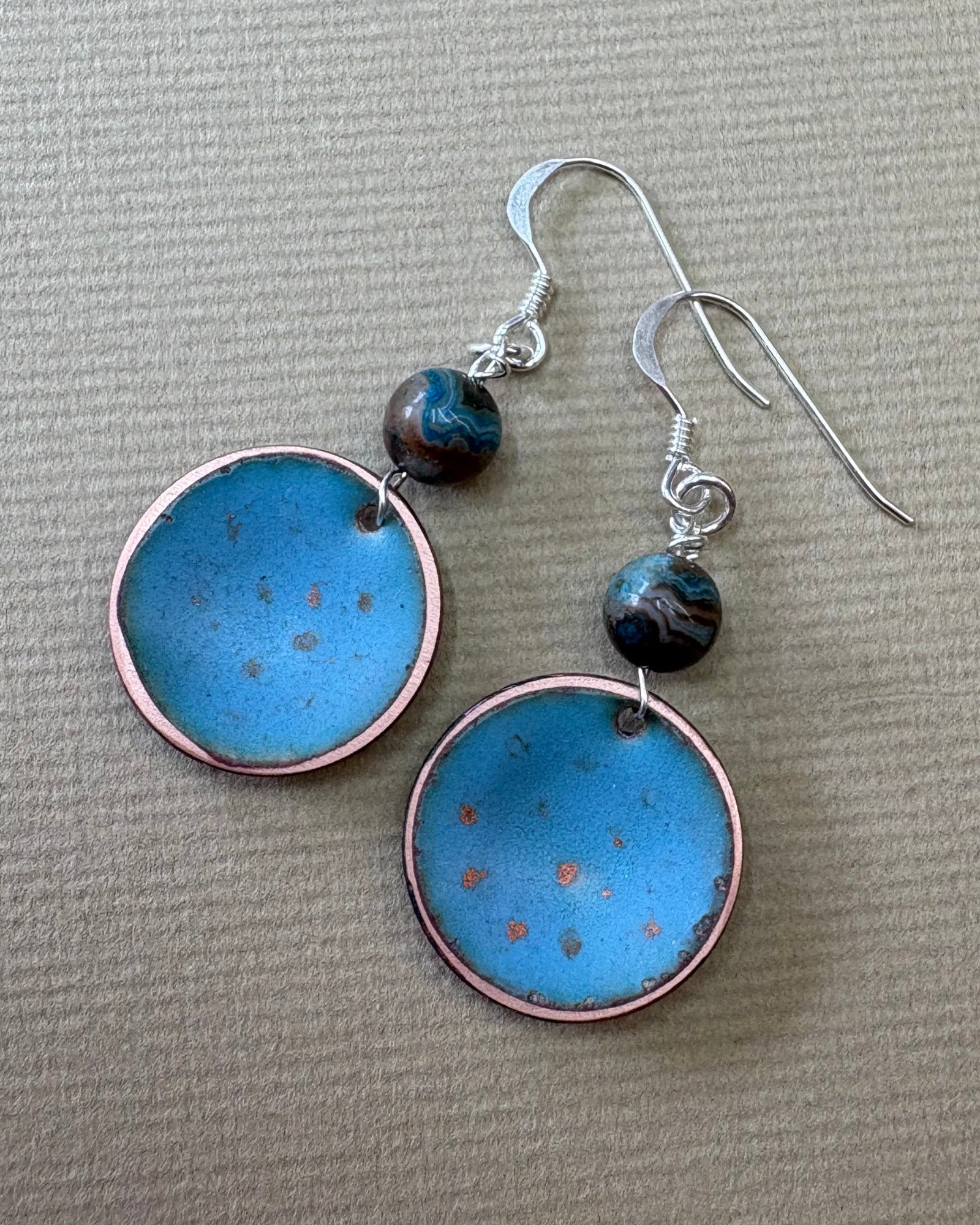 A pair of handmade dangle earrings with round light blue enamel pendants, copper-rimmed edges, and agate beads on silver fishhook wires, displayed on a pebble